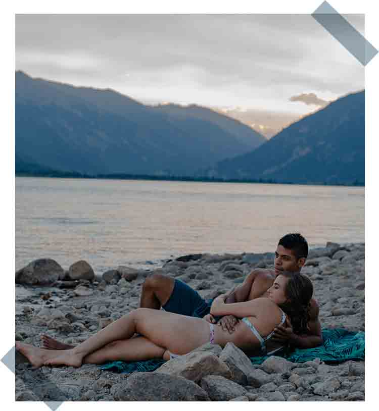 Celebrate-Again-What-To-Do-For-Your-Anniversary-diverse couple laying on a towel at the edge of an alpine lake in their swim suits snuggled up holding each other as they watch the sky fade to darkness