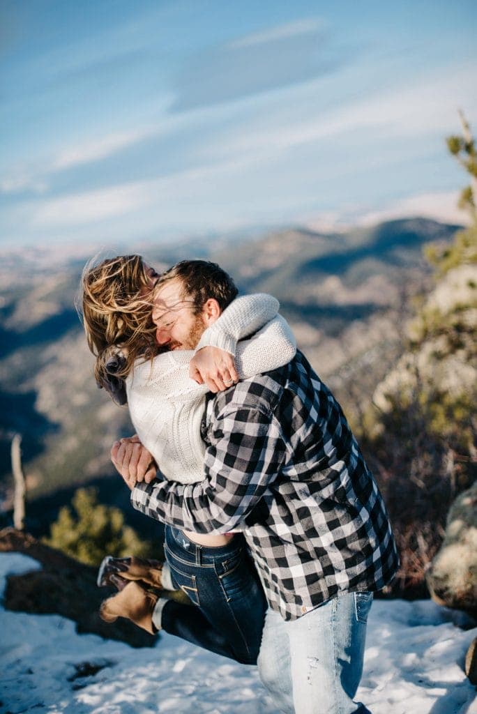 Man lifting up female spinning her around in an intimate hug on the top of a mountain top near Boulder, Colorado with sweeping mountain views and blue skies in the background
