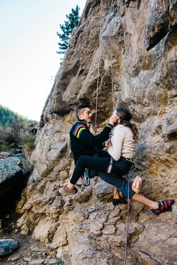 couple rock climbing both swinging from the rope as the husband wipes something off his wife's face adorably on their best anniversary trip ideas
