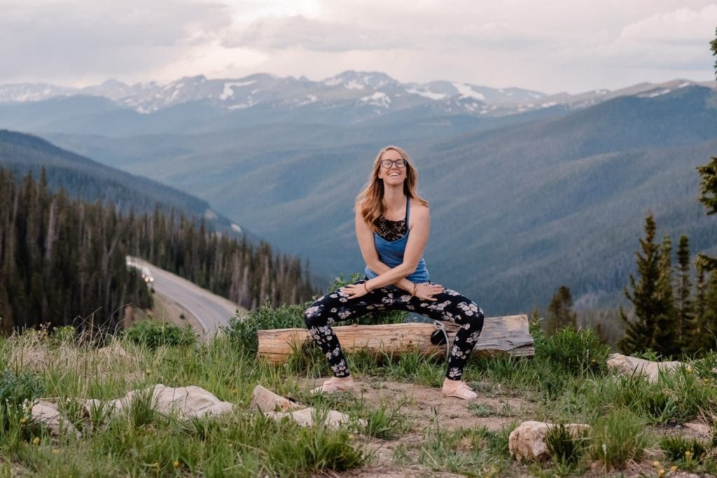 emmy standing on mountain top view point squatting with her arms crossed smiling at mountain with wild flowers in front of her on the ground and a sweeping mountain view in the background