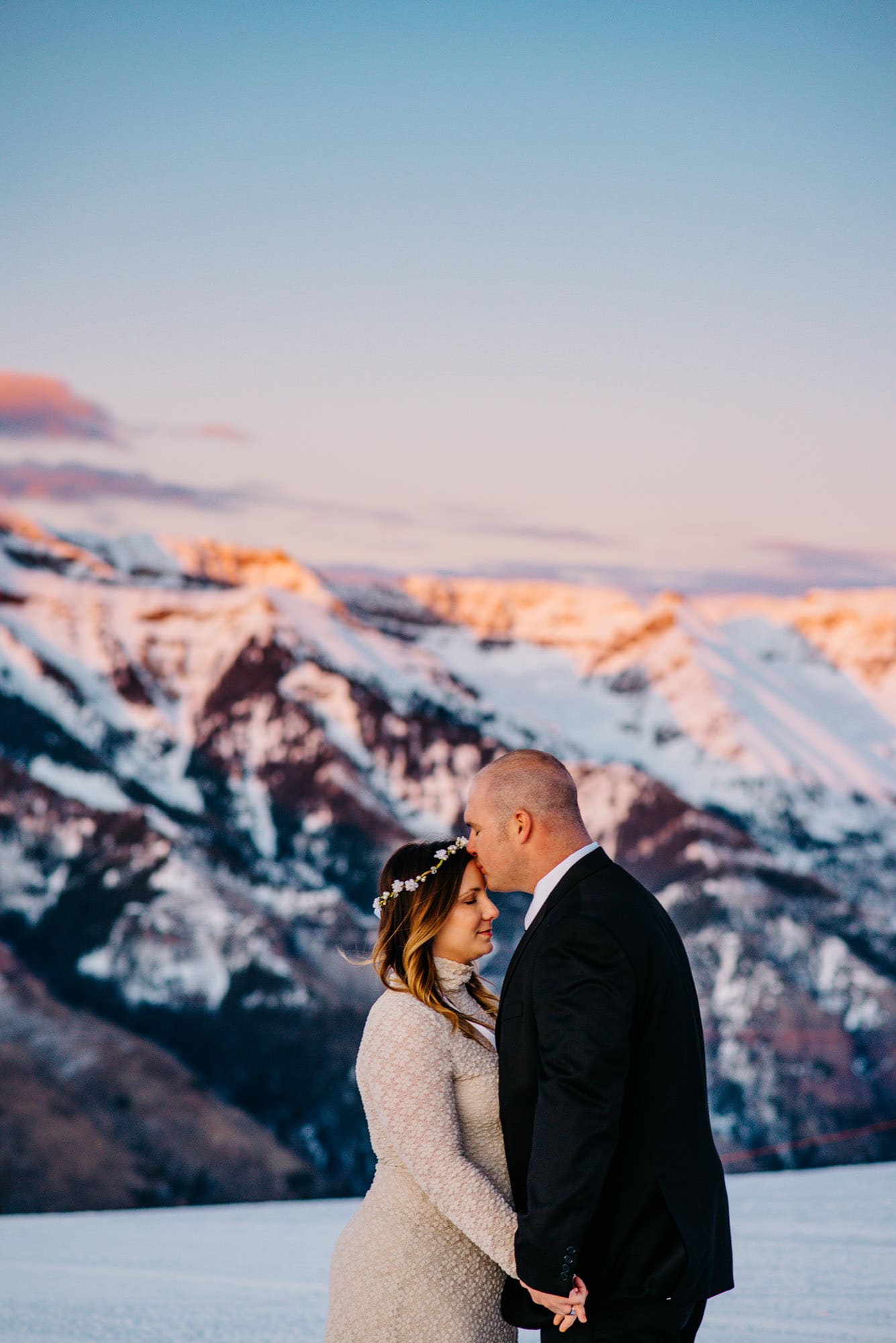 groom kissing brides head on top of a mountain in the snow as the sun glows behind them on the mountains