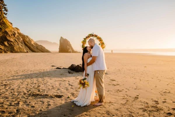 couple standing on beaching having a cannon beach elopement as the sunsets behind them with sea rocks in the distance