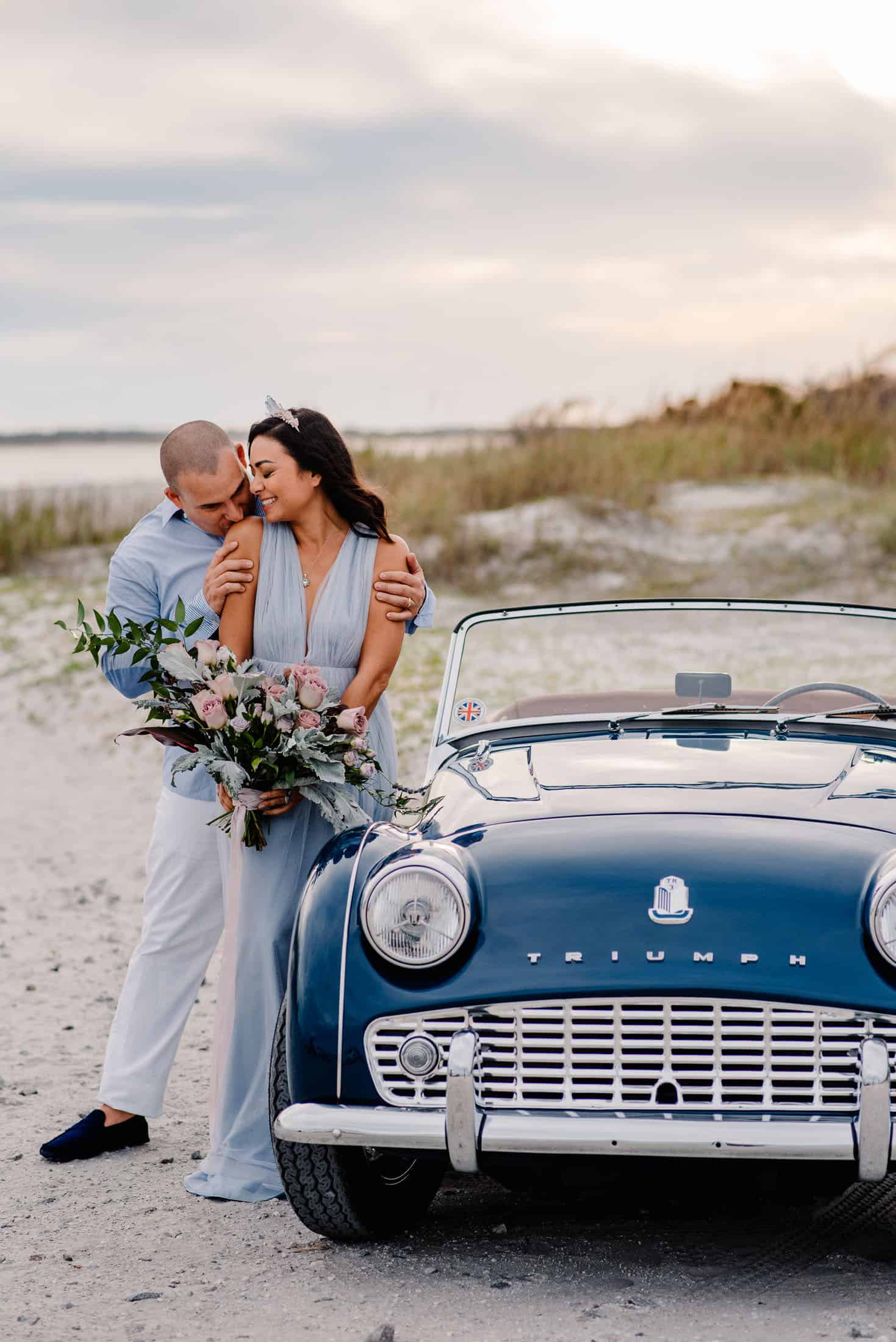 couple with a vintage car having a Cannon Beach elopement at sunset. Groom is kissing bride on the shoulder and she's turning her head towards him