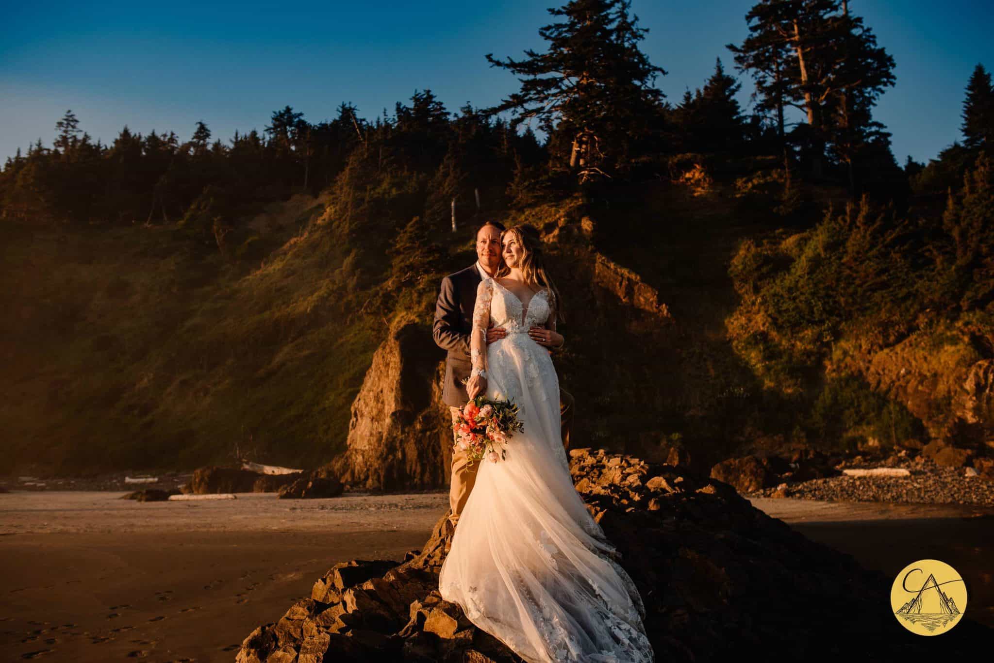 oregon elopement photographer taking photo of couple standing on the oregon coast as the sun shines on them and the cliffs are behind them