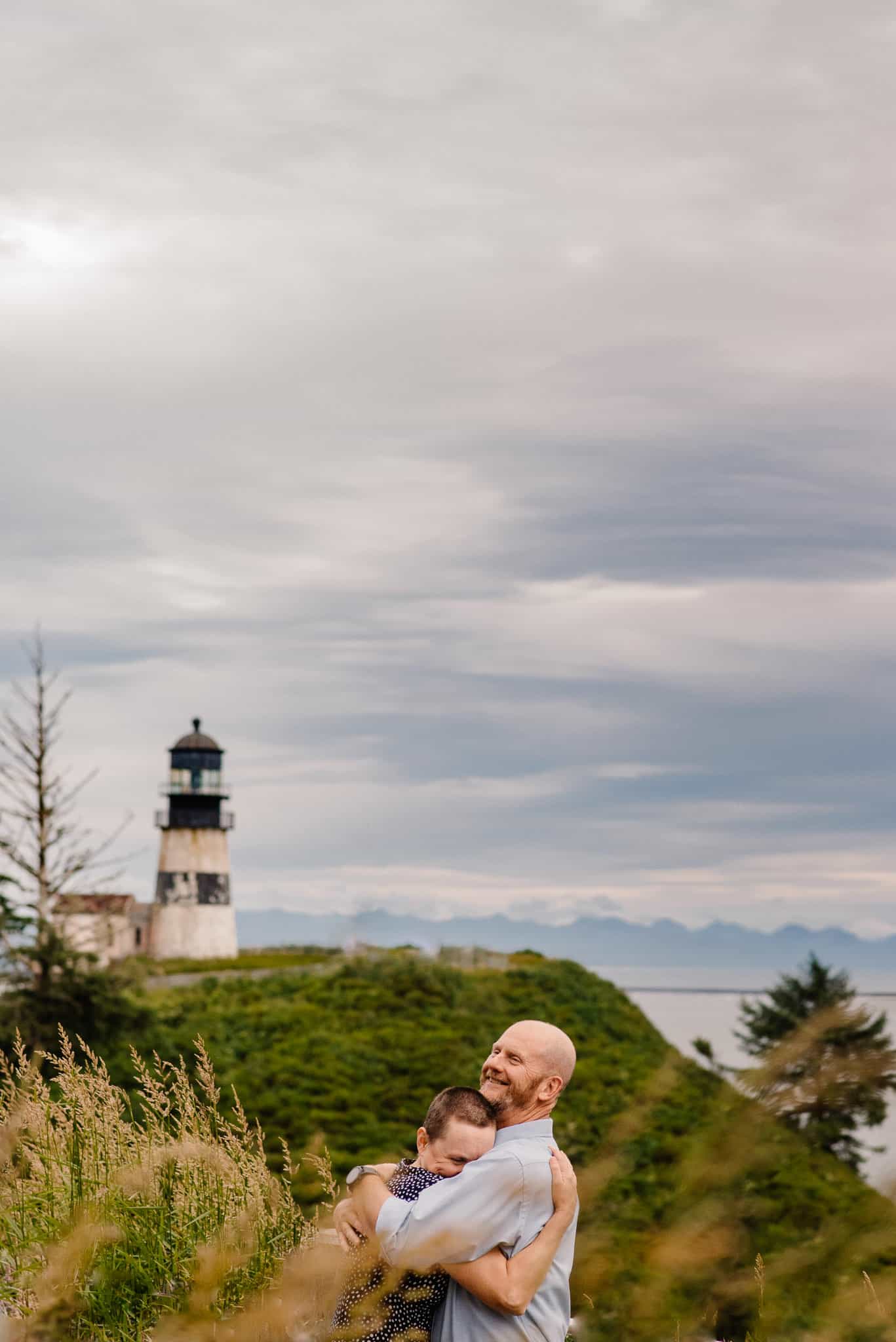 couples hugging near the ocean with a lighthouse in the background