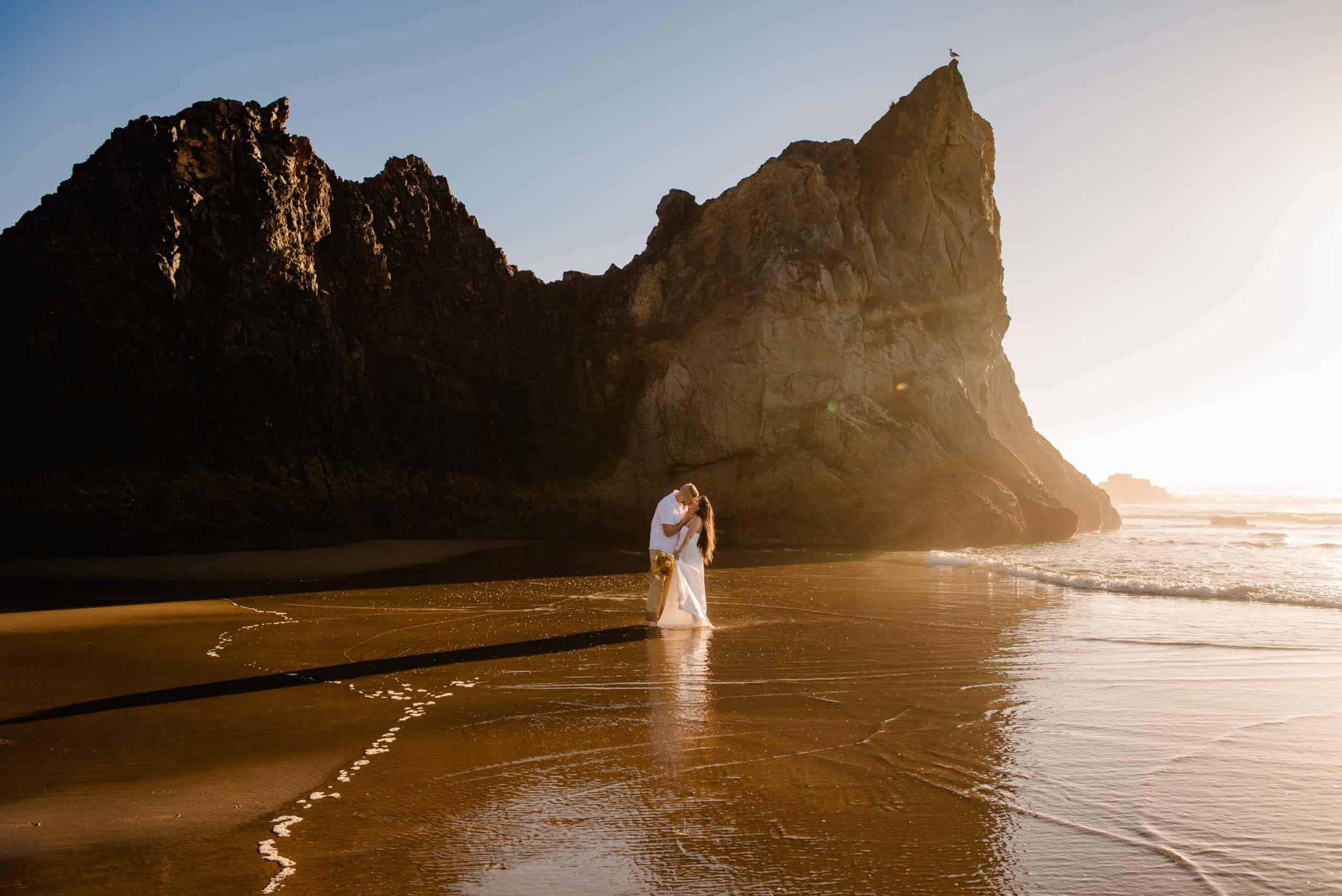 couple off in the distance with a sea rock behind them and water coming up as their oregon elopement photographer take their pictures