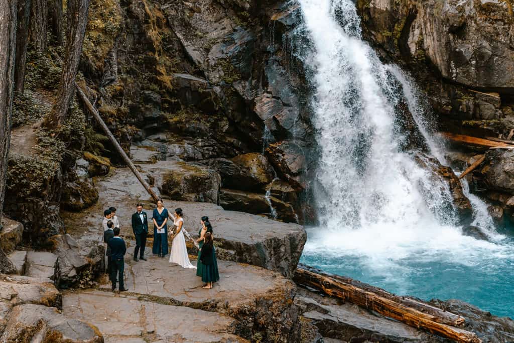 Couple in wedding attire gather cliffside with their small wedding party as a dramatic turquoise waterfall makes for the perfect backdrop for this waterfall wedding