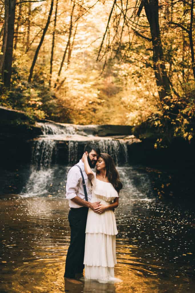 couple standing in the river with a waterfall behind them photo by Sarah Katherine Davis Photography