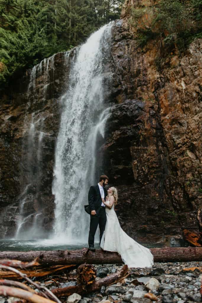 Bride and groom embrace infant of a gushing waterfall in their wedding attire after learning how to elope in Oregon