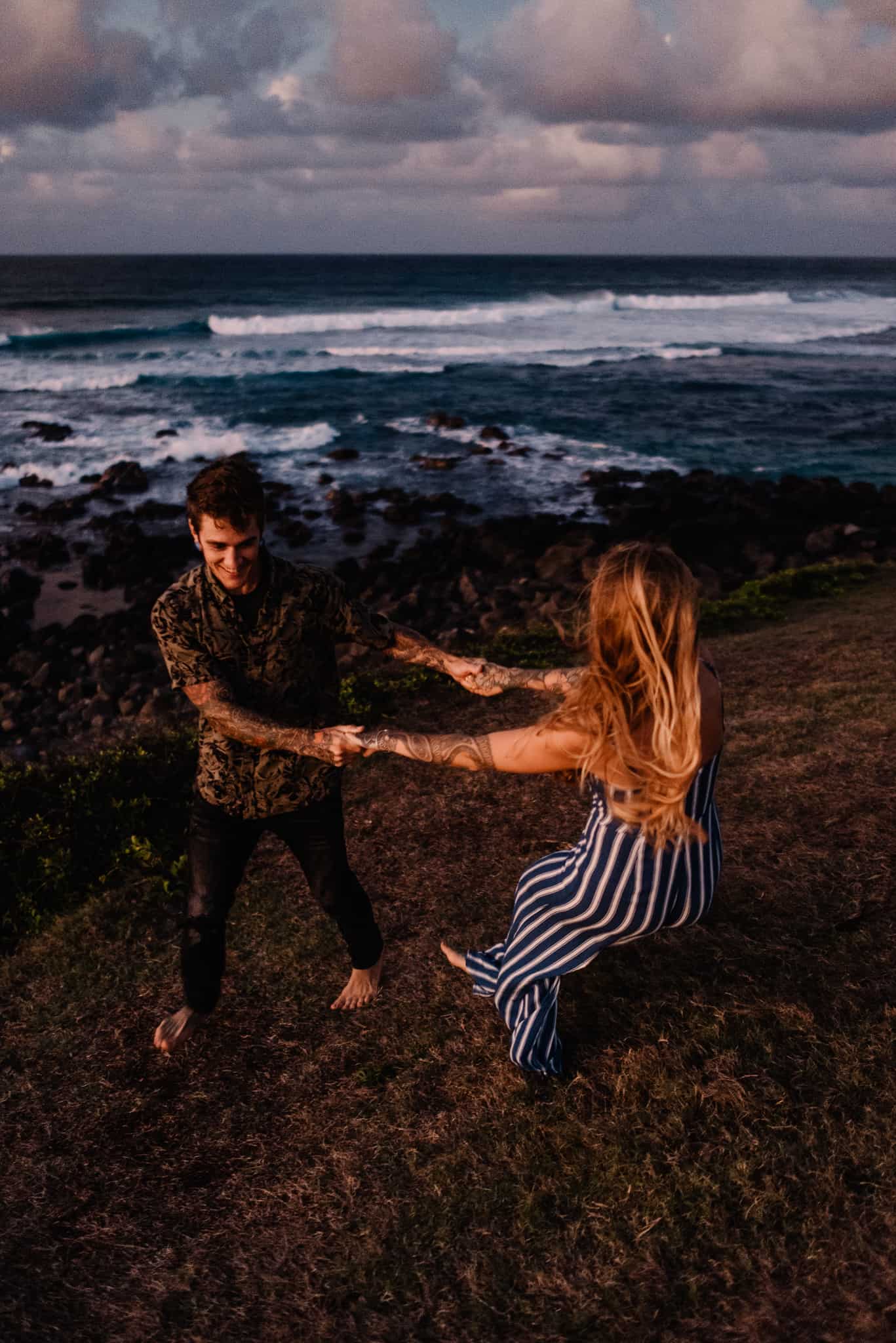 couple holding hands spinning with the sun turning the ocean and clouds vibrant pinks and purples on the ocean as their Hawaii elopement photographer takes their photo