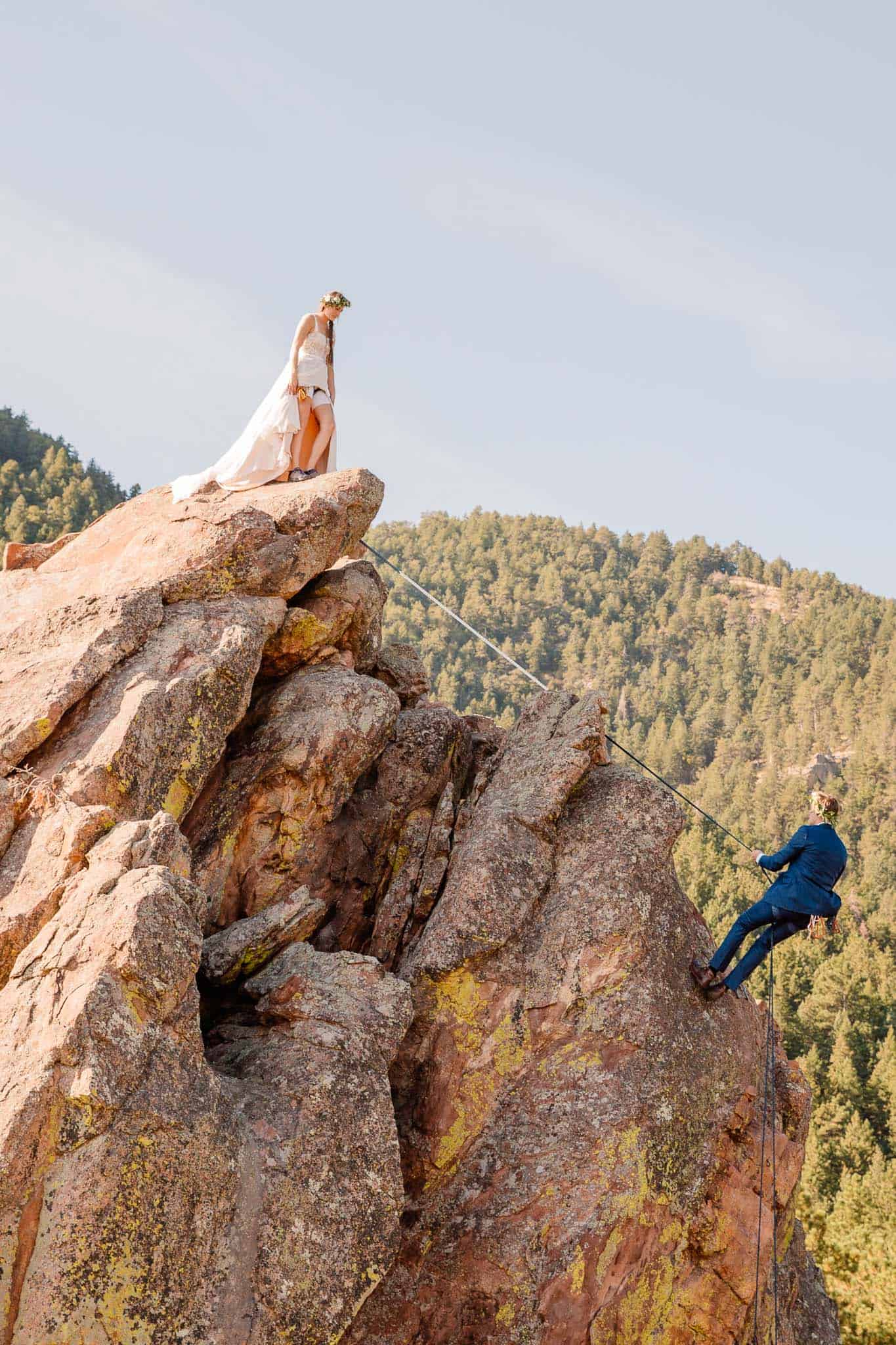 bride and groom rock climbing as bride watches groom rappel down a mountain