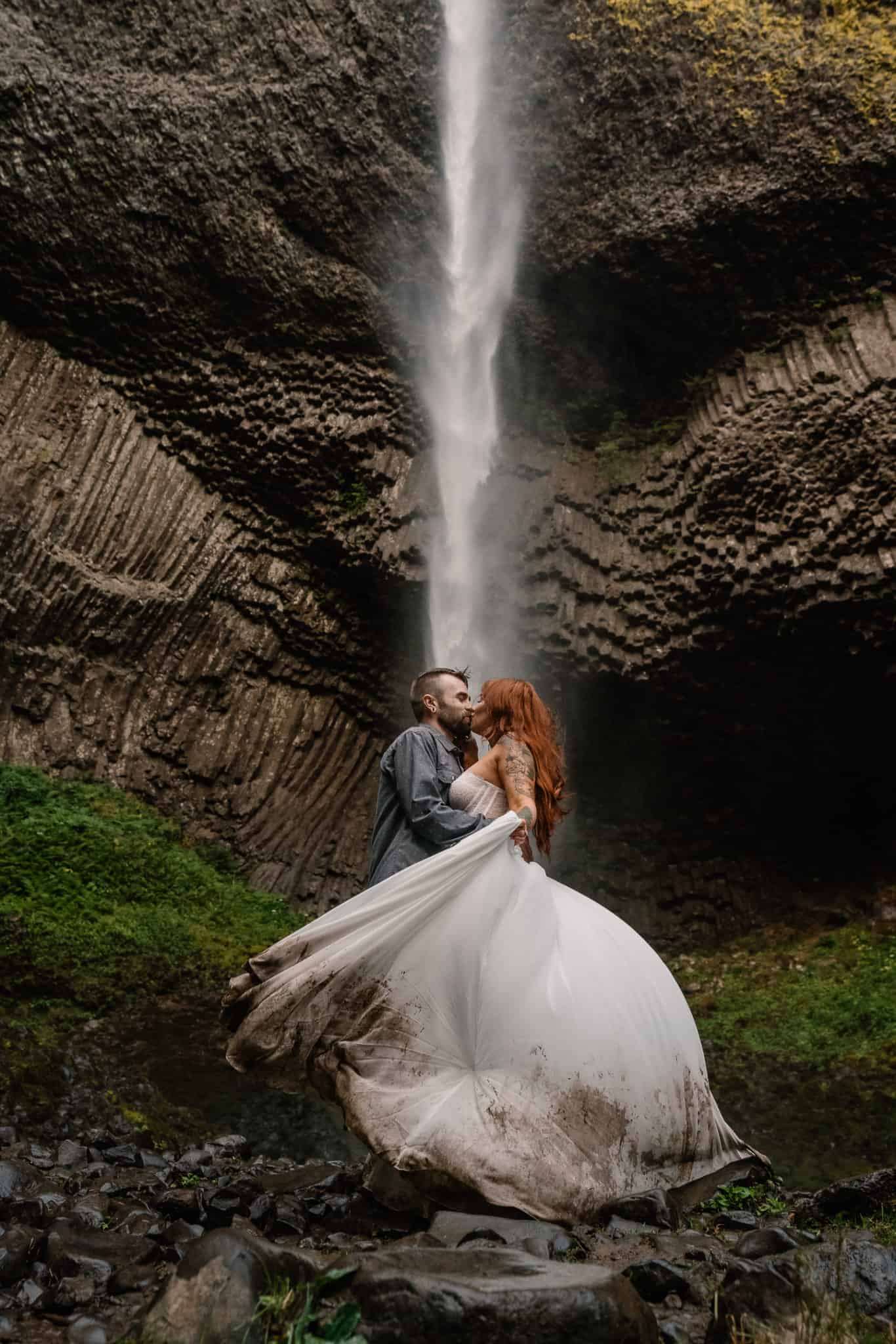 Bride twirls her muddy wedding dress as she kisses her groom with a plunging waterfall in the background making this one of the best places to elope in Oregon