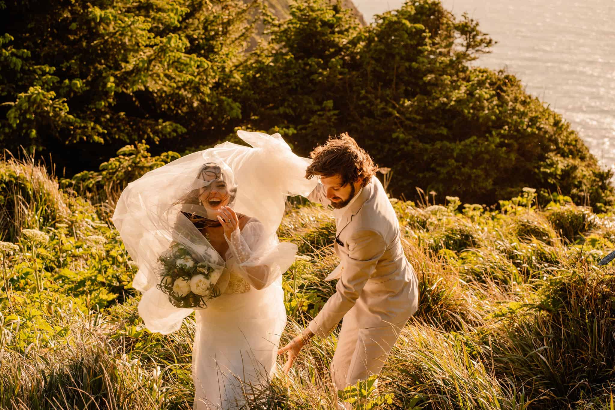 Bride and groom walk through a coastal meadow with golden sunlight behind them and high winds blowing through their wedding attire