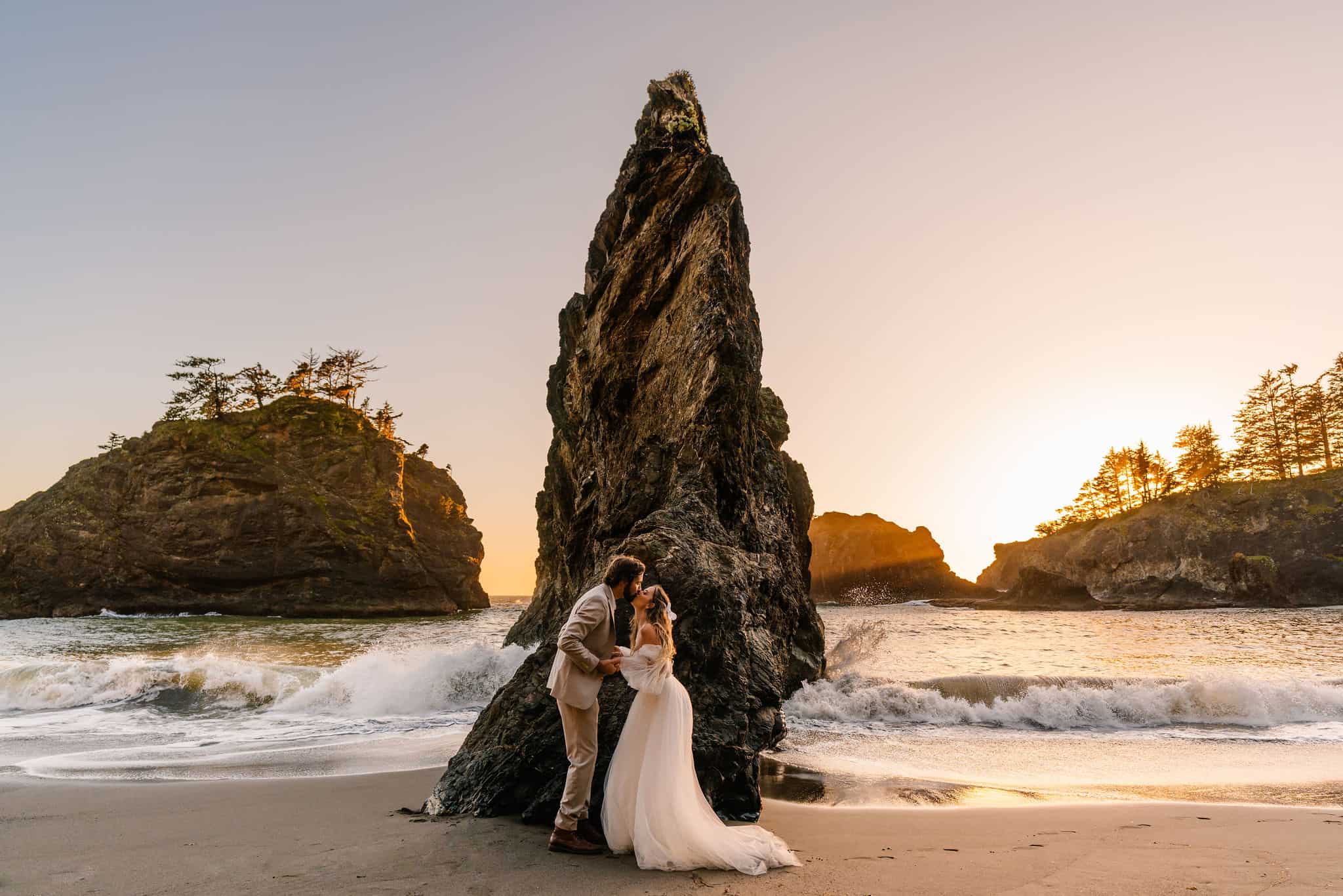After learning how to elope in Oregon bride and groom embrace in front of a jagged sea rock as the sun sets behind them
