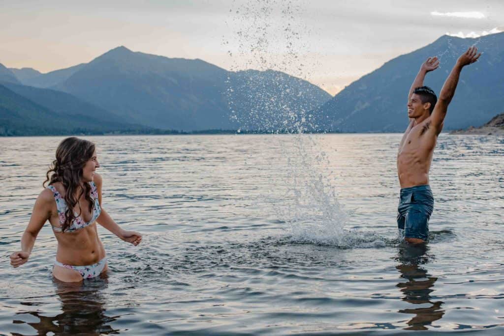 couple splashing water in front of each other in an alpine lake in their swimsuits during a couples connection experience