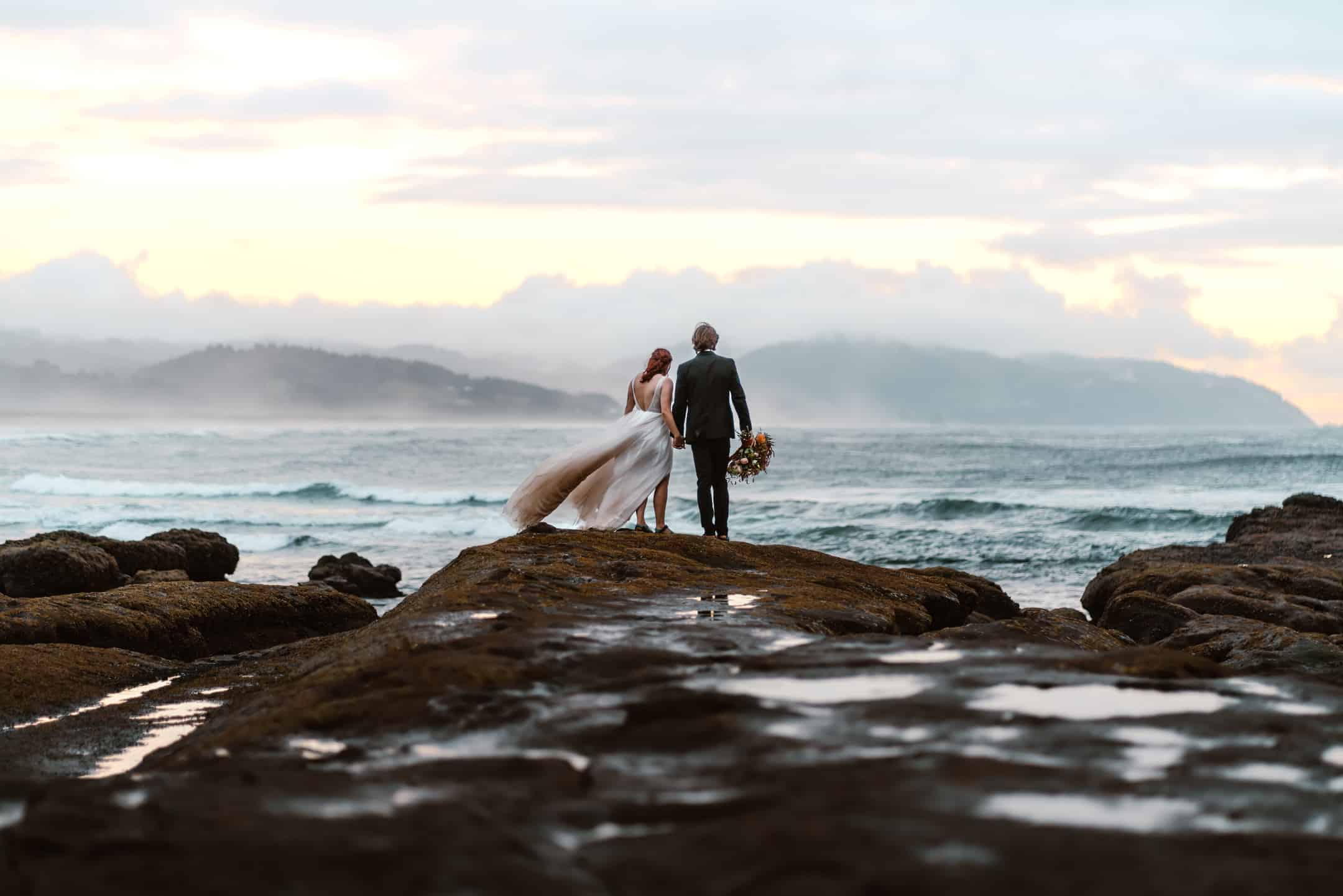 Couple holds hands in their wedding attire, framed by tide pools as their with waves crashing behind them as their Oregon elopement photographer takes their photo