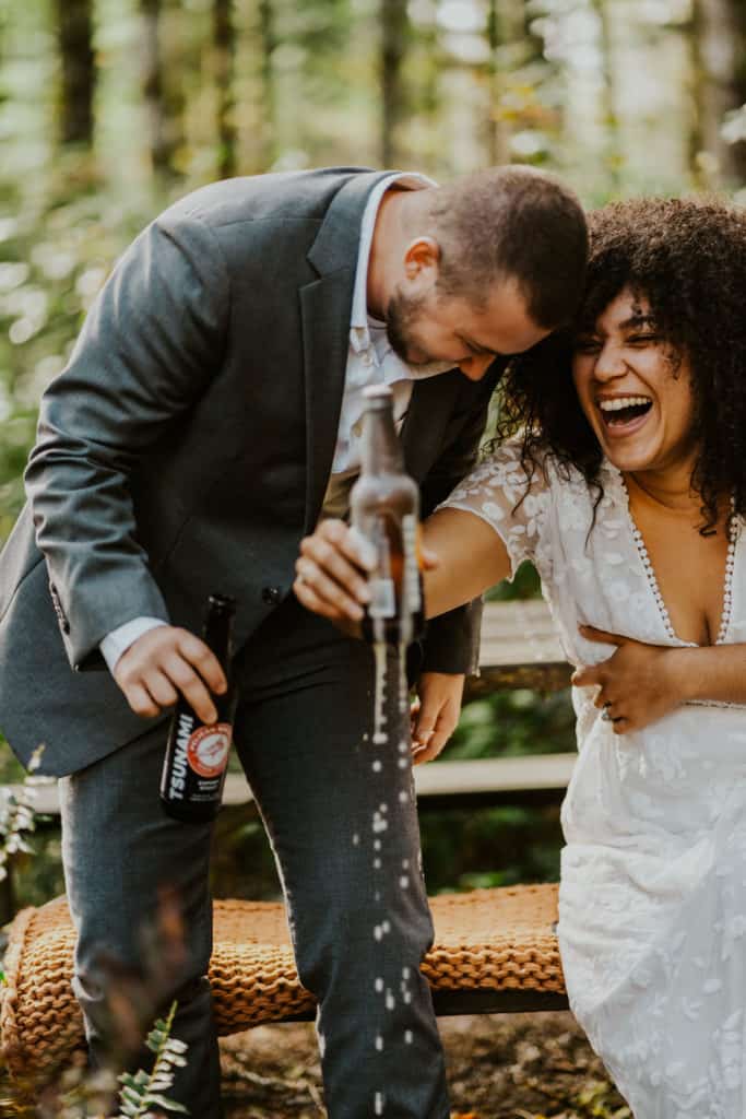 couple holding drinks in the forest laughing as their Oregon elopement photographer takes their photo