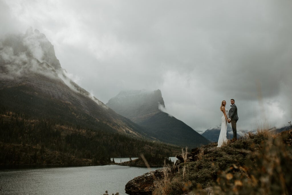A couple stands in their wedding attire on a cliff in one of the best places to elope, Montana. Moody fog hangs over the mountains giving a moody vibe.