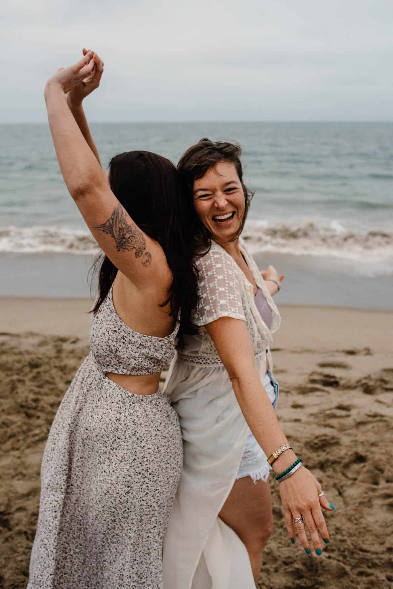 females back to back one with arms raised the other arms out on a beach during a group boudoir self love photography experience