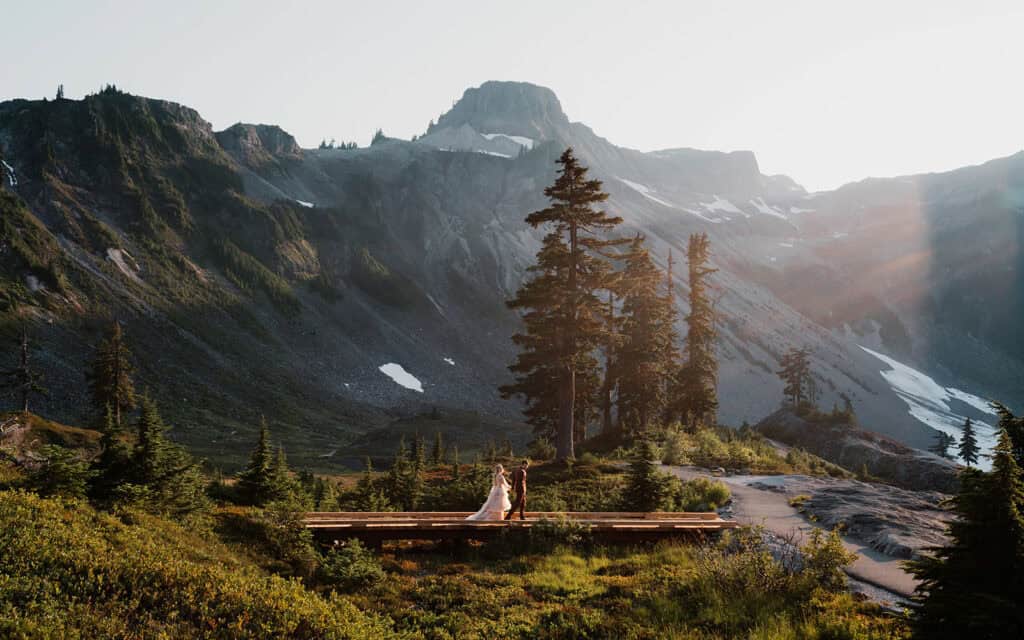 a couple in wedding attire walks on a wooden bridge with mountains and tall trees behind them. A lens flare shines on the right side of the wide, landscape shot taken during their mount baker elopement, one of the best places to elope