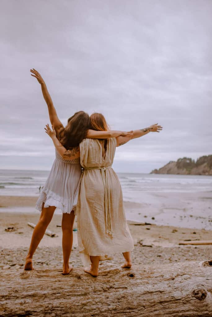 woman holding arms on a log by the beach as they heal the sister wound during a women's retreat