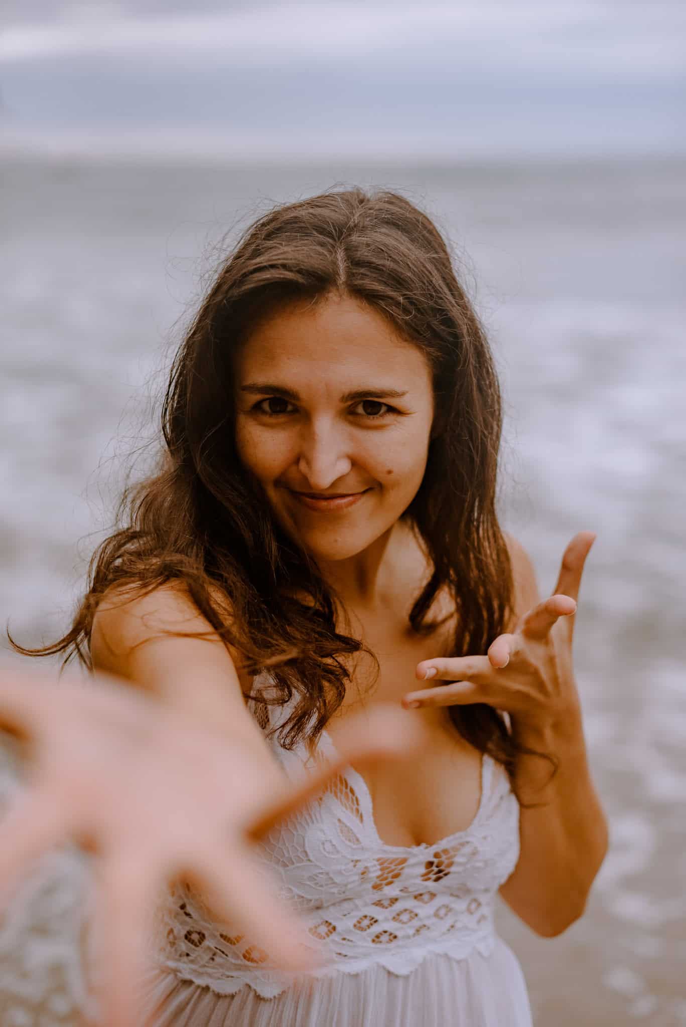 woman smirking reaching her hands out toward you with the ocean in the background