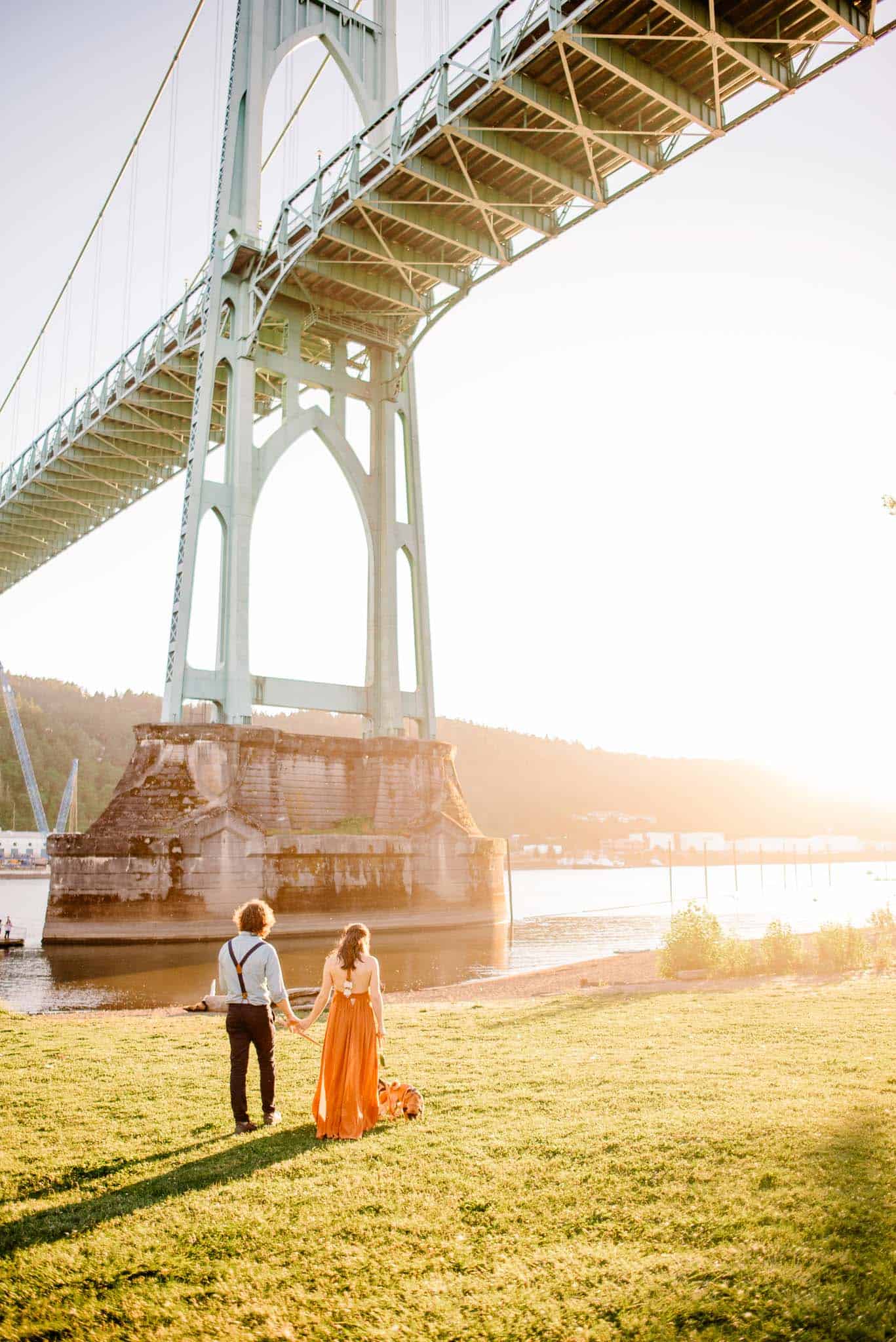 couple walking on grass near the rive under Cathedral Park under St. John's bridge during engagement photos in Cathedral Park