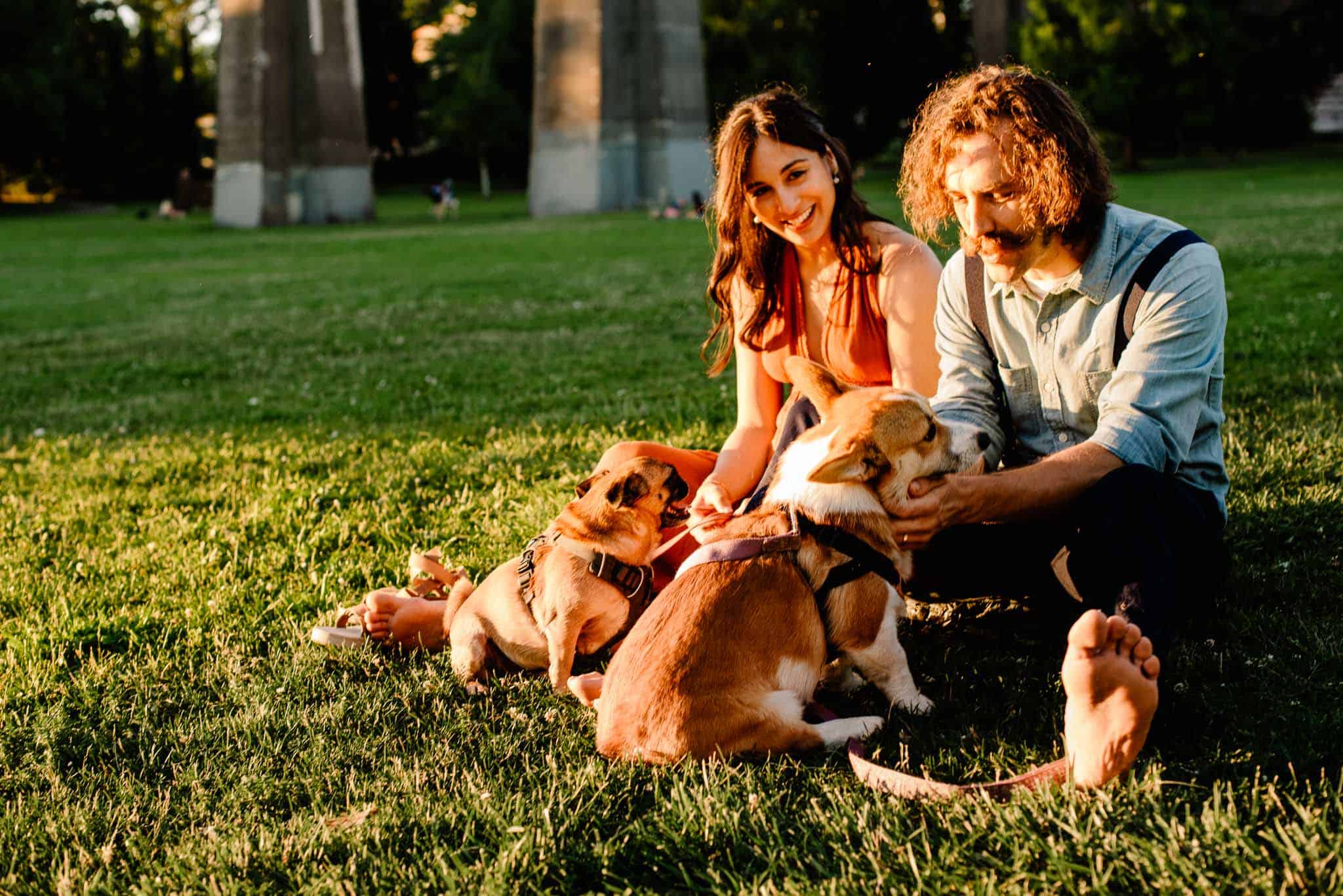 couple sitting on grass playing with their dogs in Cathedral Park during engagment photos