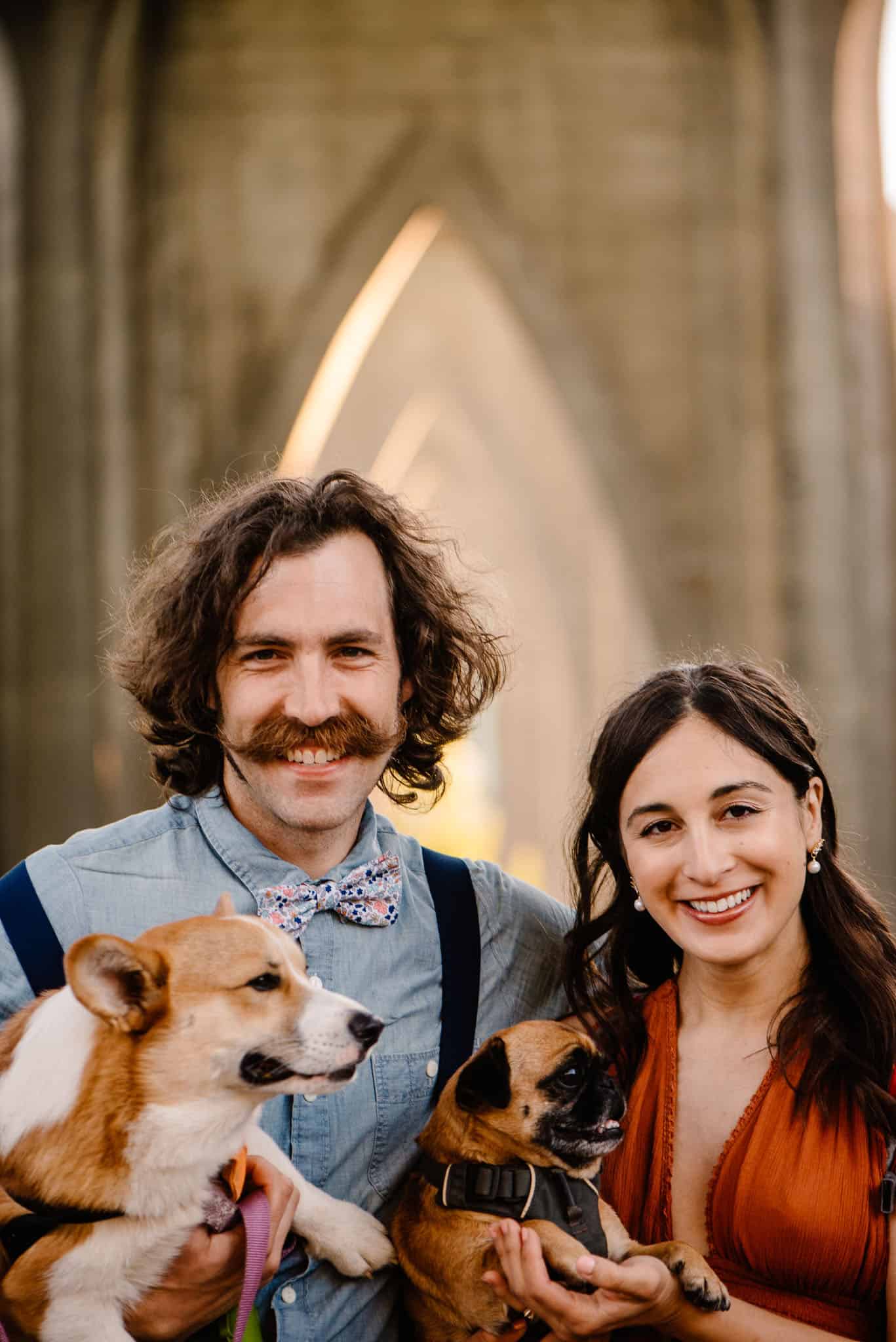 Couple smiling holding dogs with Cathedral Park arches behind them at susnet The arches: We know Cathedral Park’s arches, the best place for engagement photos in Portland, Oregon. 